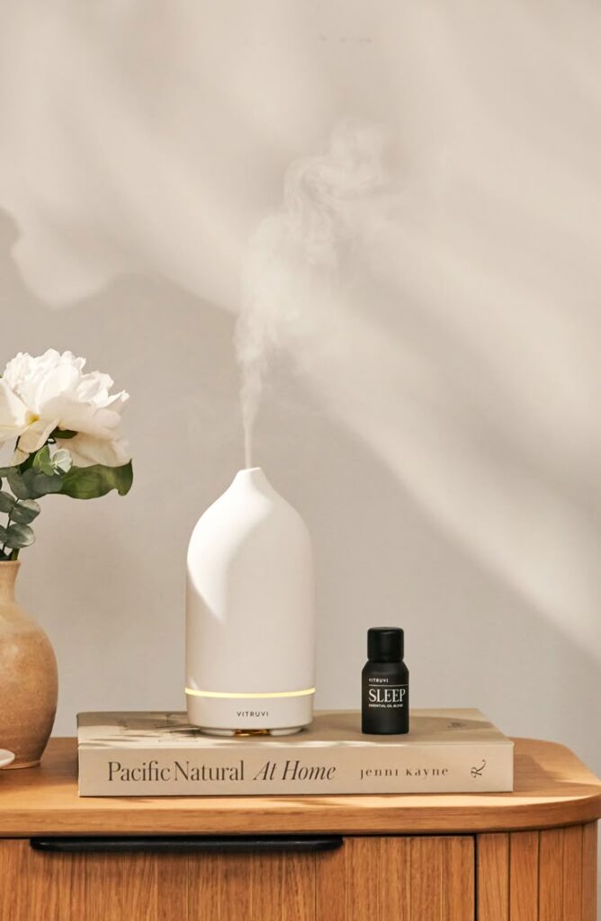 Aromatherapy diffuser emitting mist next to essential oil bottle on wooden table with flowers and books.