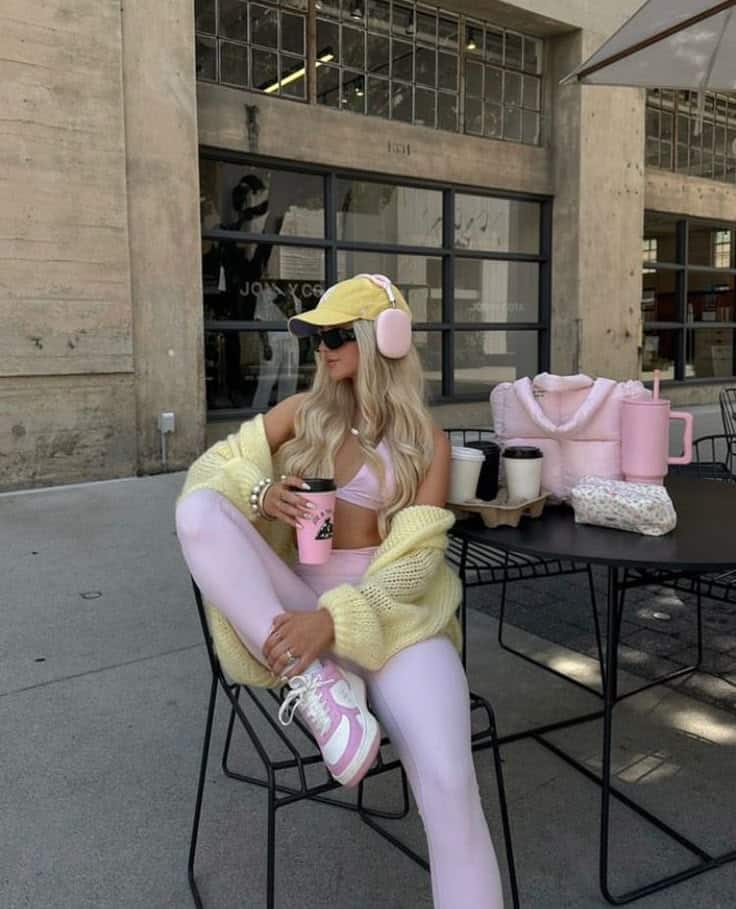 Woman in pastel activewear and headphones, sipping coffee outdoors, surrounded by pink accessories.