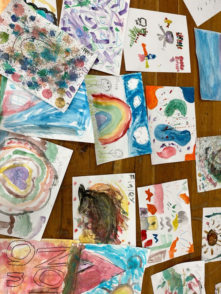 Colorful children's artwork spread across a wooden floor, showcasing various creative paintings and drawings.