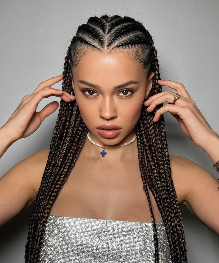 Young woman with braided cornrows, wearing a sparkly top and a necklace with a blue cross charm, posing confidently.