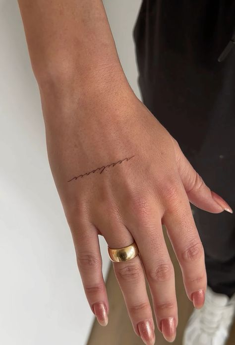 Close-up of a hand with minimalist script tattoo on knuckle, wearing a gold ring, showcasing trendy nail art.