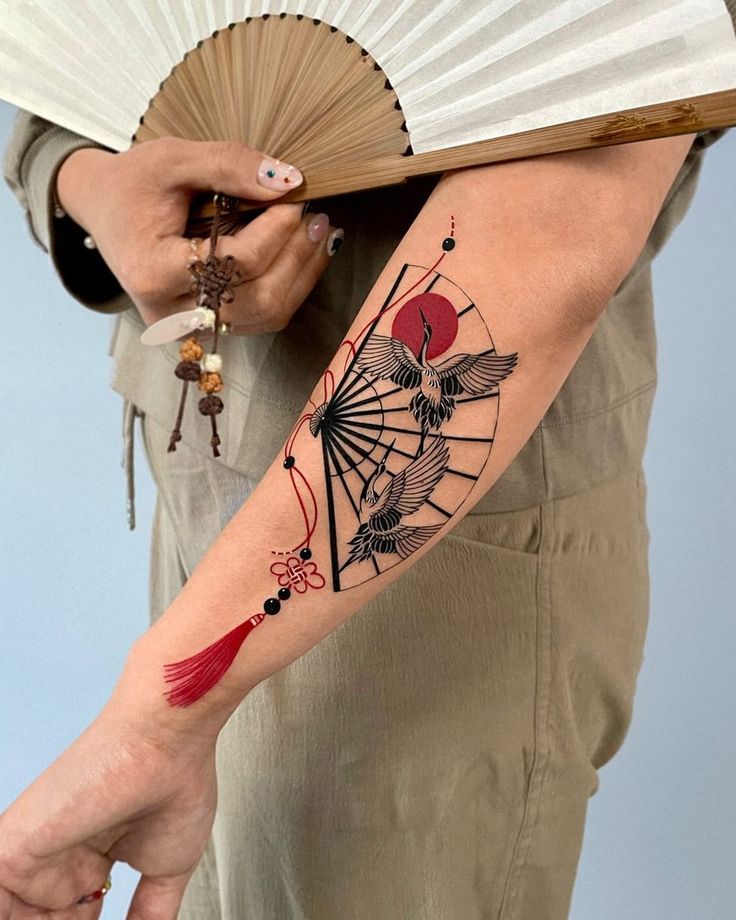 Elegant forearm tattoo of a crane with red sun and fan, complemented by a matching Japanese-style fan accessory.