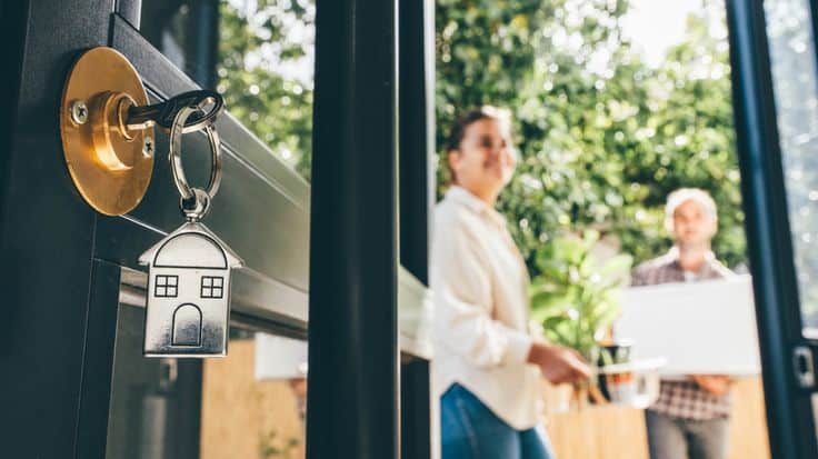 House key in door as couple moves in, carrying boxes and plants, symbolizing new home and fresh beginnings.