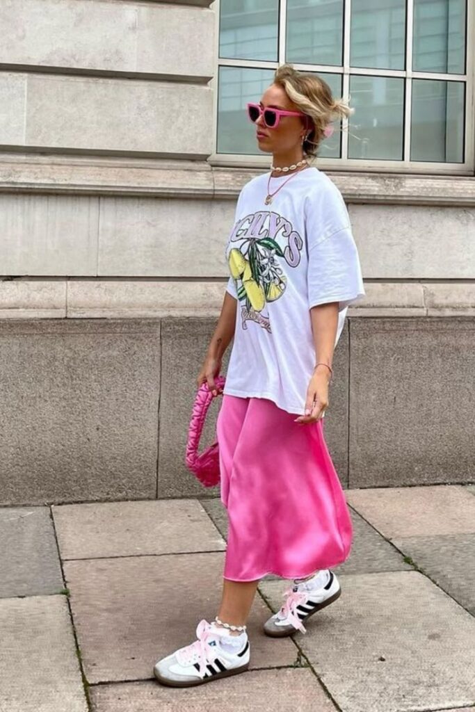 Fashionable person in pink shades, graphic tee, pink skirt, and sneakers walking on city sidewalk with trendy pink bag.