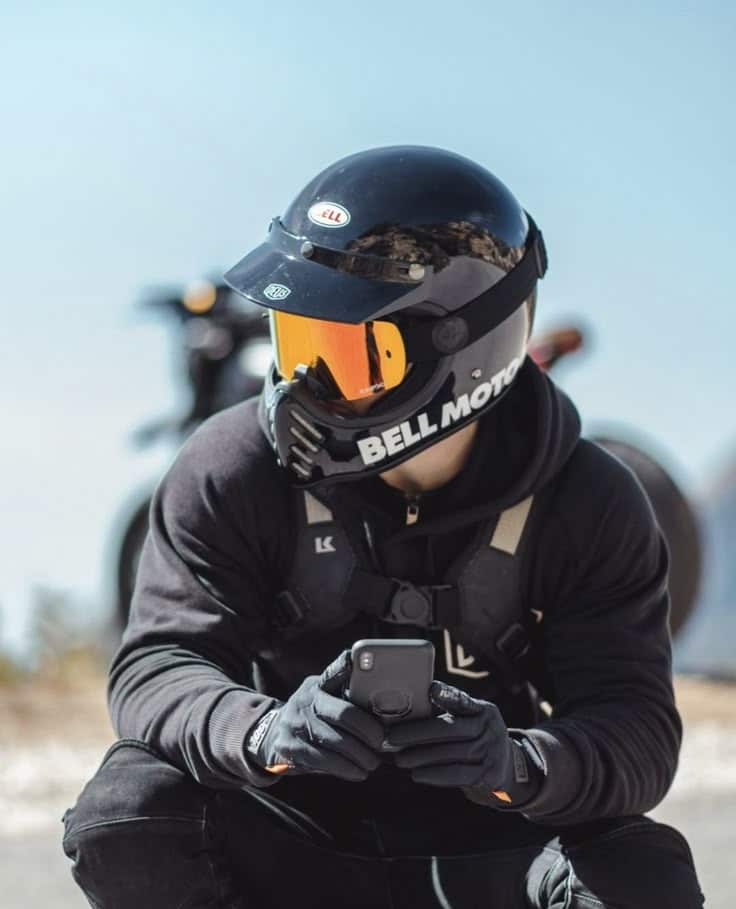 Motorcyclist in black gear and helmet checking phone, with a bike in the background under a clear blue sky.
