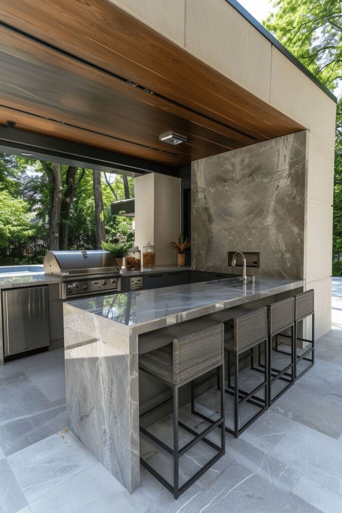 Modern outdoor kitchen with marble countertop, grill, and bar stools, surrounded by lush greenery.