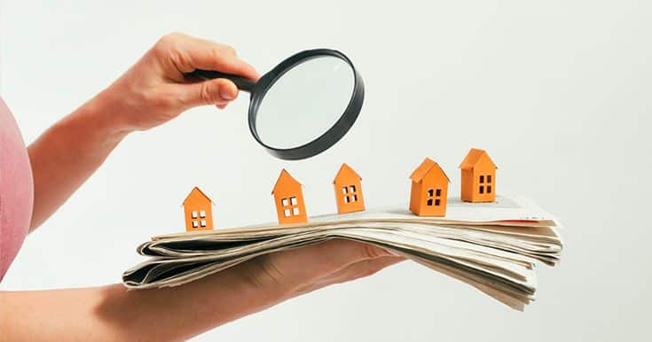 Person holding a magnifying glass over small house models on a newspaper, representing real estate market analysis.