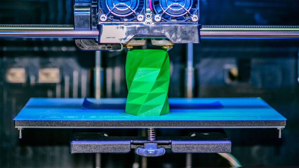 3D printer creating a green geometric object on a blue build plate in a close-up view.