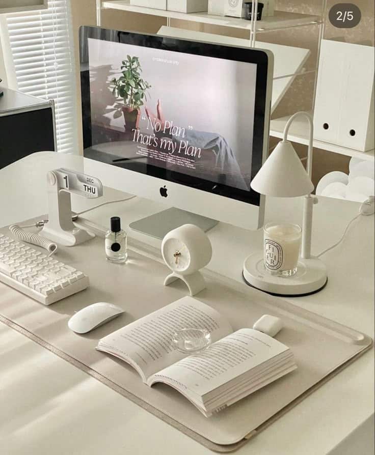 Minimalist white desk setup with computer, keyboard, open book, lamp, and decor items for productivity and style.