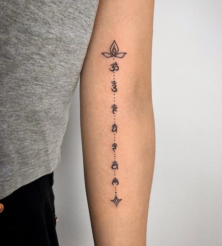 Minimalist Sanskrit tattoo with symbols on forearm, featuring a lotus design and dots, inked in black.