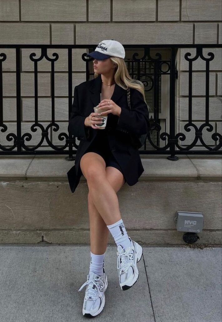 Fashionable woman in blazer, cap, and sneakers sitting on a bench, holding a drink; chic urban style.