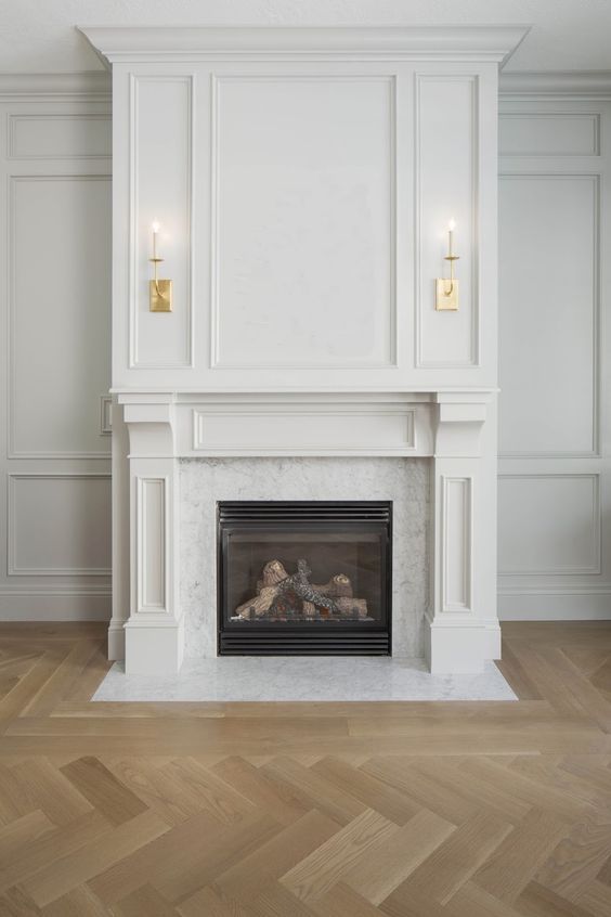 Elegant white fireplace with gold sconces, marble detailing, and herringbone wooden floor in a classic living room.