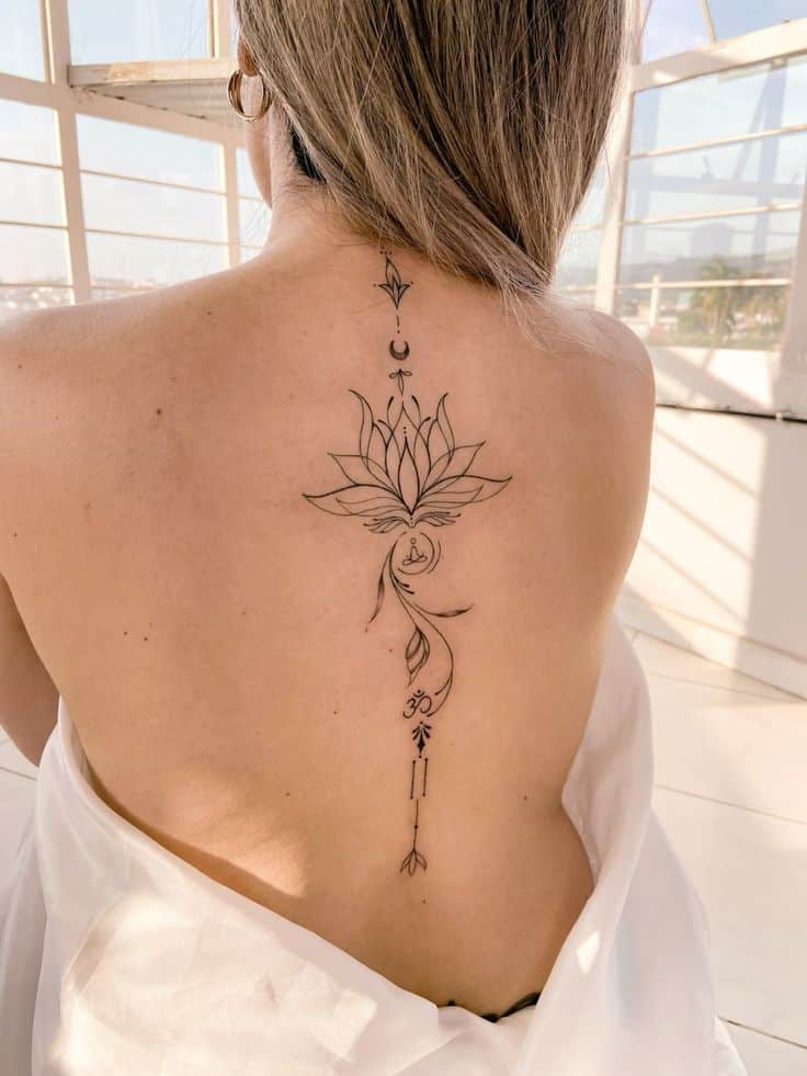 Back view of woman with intricate lotus tattoo down spine, wearing a draped white cloth, in a bright room.