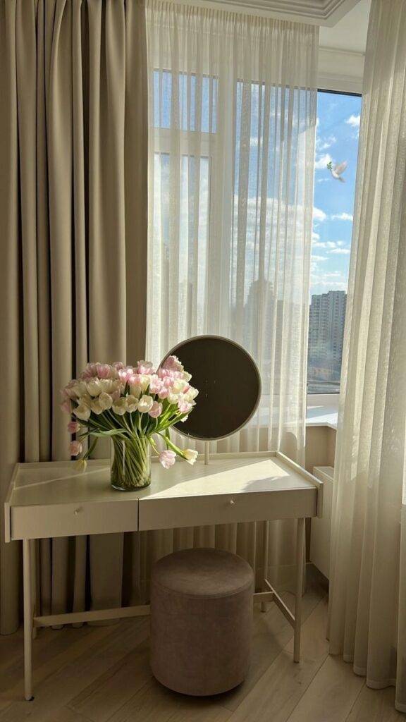 Vanity table with pink and white tulips by a bright window, sheer curtains, and a round mirror.