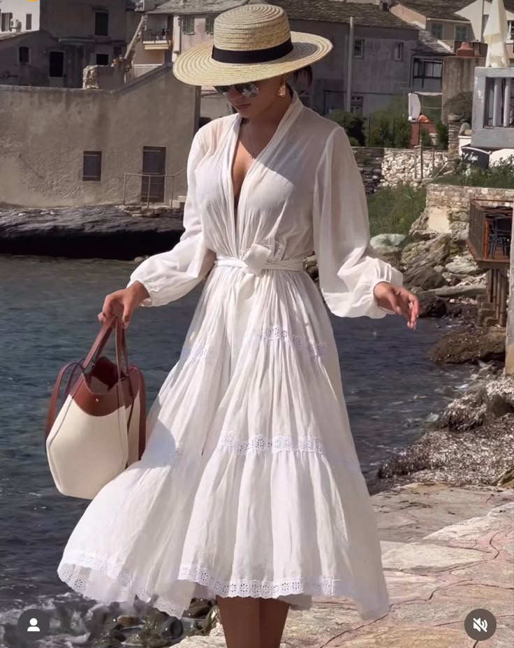 Woman in flowing white dress and straw hat walking by seaside village.