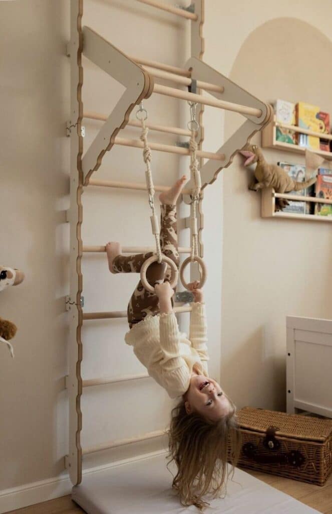 Child playing on indoor gym rings, hanging upside down with joy in a cozy room with toys.