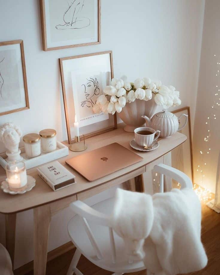 Cozy home office with laptop, candles, art, flowers, and tea on a wooden desk in a softly lit room.