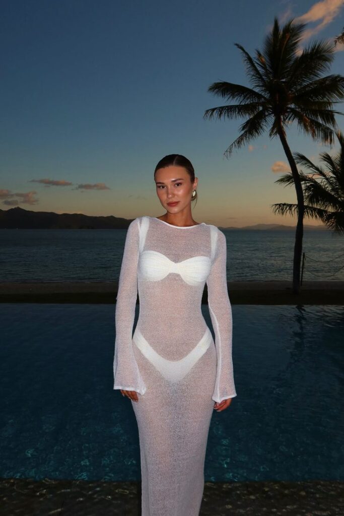 Woman in sheer white dress poses by ocean at sunset, palm tree and mountains in background.