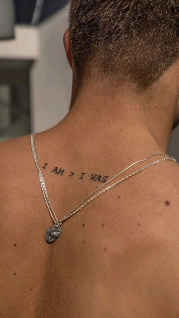 Back tattoo saying I AM > I WAS with silver necklace and coin pendant.