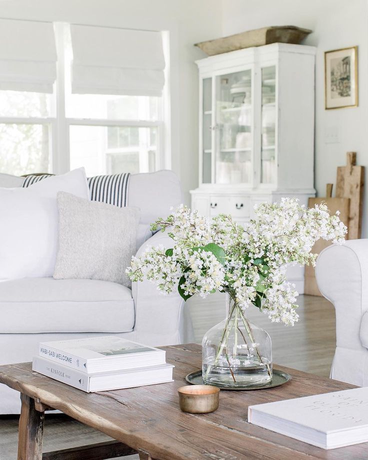 Bright living room with white sofa, rustic coffee table, glass vase of white flowers, and decorative books.