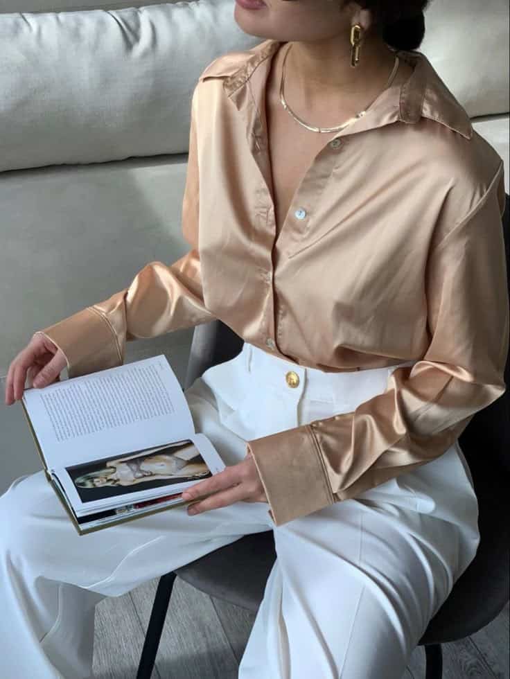 Woman in a satin blouse reads a fashion magazine, sitting in a chair, wearing white pants and accessorized with gold jewelry.