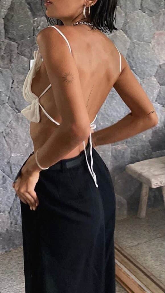 Fashionable woman in a backless top and black pants with a sun tattoo, standing against a stone wall background.