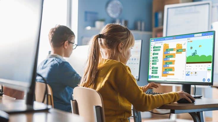 Children in a classroom learning programming on computers, engaging with a visual coding interface.