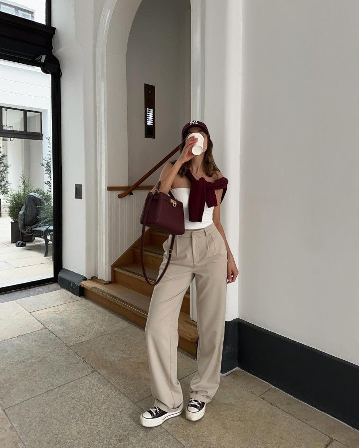 _ 71 Casual chic outfit with white top, beige pants, and burgundy accessories, holding coffee in a stylish interior setting. | Sky Rye Design Casual chic outfit with white top, beige pants, and burgundy accessories, holding coffee in a stylish interior setting.
