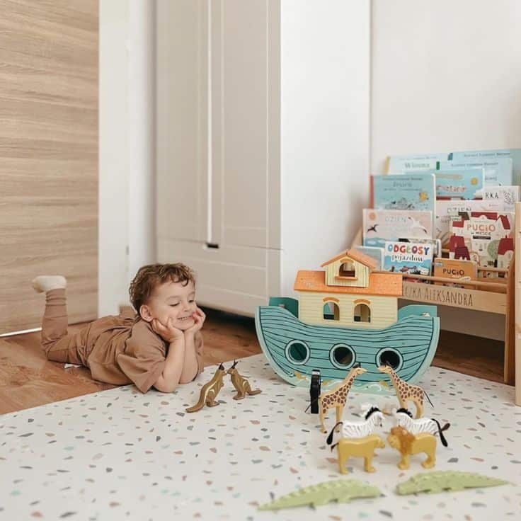Child playing on floor with wooden animal toys and ark, surrounded by books on shelf in cozy room.