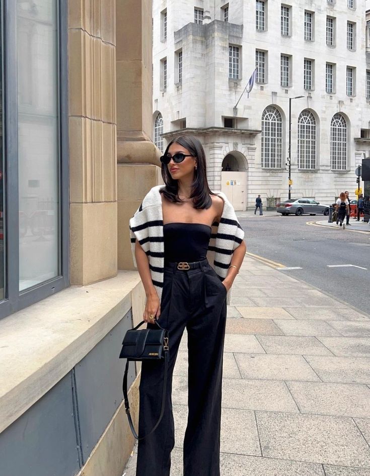 _ 72 Stylish woman in sunglasses wearing black outfit with striped sweater, holding a handbag in urban street setting. | Sky Rye Design Stylish woman in sunglasses wearing black outfit with striped sweater, holding a handbag in urban street setting.