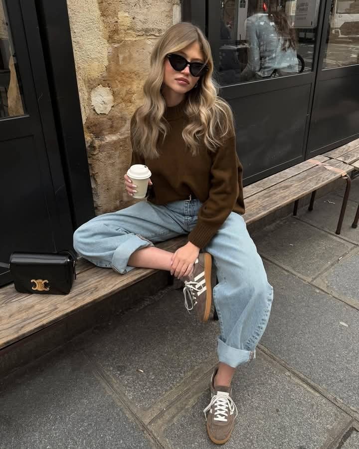 Woman in casual outfit with coffee, sitting on a bench wearing sunglasses, brown top, and jeans.