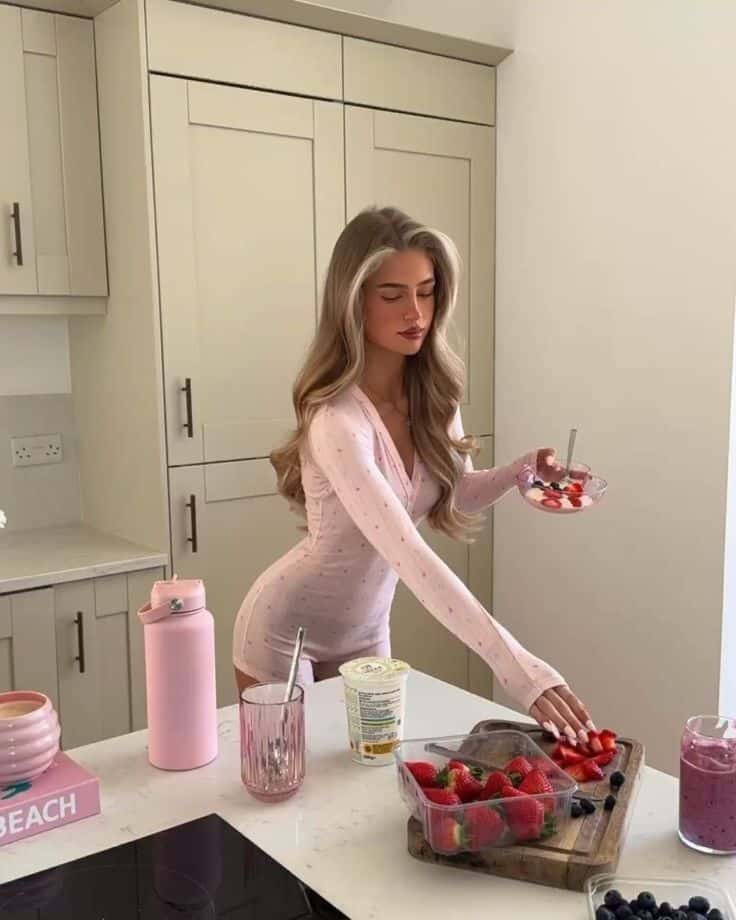 Woman in pink preparing a fruit bowl in a modern kitchen with strawberries, yogurt, and smoothie on the counter.