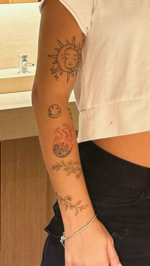 Person's arm with a sun, moon, smiley face, and floral tattoos in a casual indoor setting.