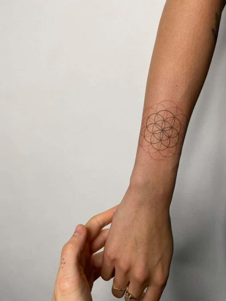 Geometric flower tattoo on forearm, hand with rings gently touching, minimalistic ink design.