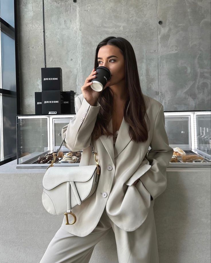 Woman in a beige suit drinking coffee, holding a stylish white purse. Urban café setting with modern decor.
