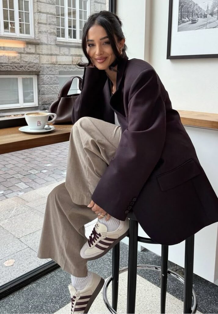 Fashionable woman in oversized blazer and sneakers sits at a cafe, enjoying coffee by a window.