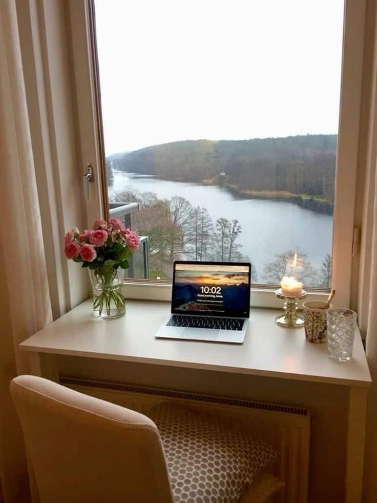 Cozy home office setup with laptop, candle, and flowers by a window overlooking a scenic river and forest view.