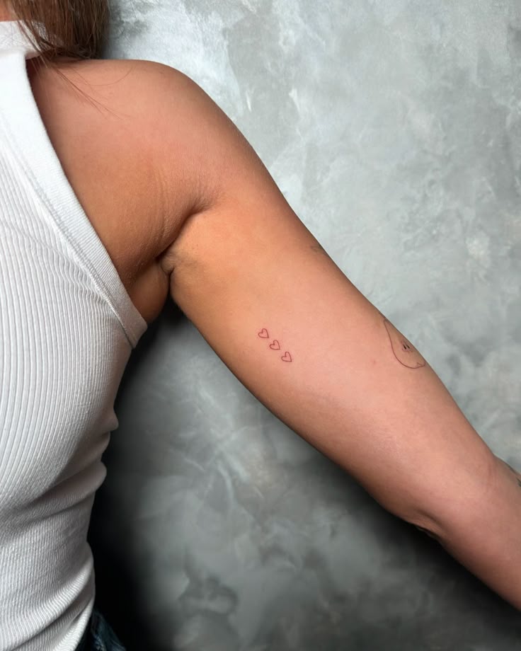 Close-up of a woman's arm with a small heart tattoo design, wearing a white sleeveless top.