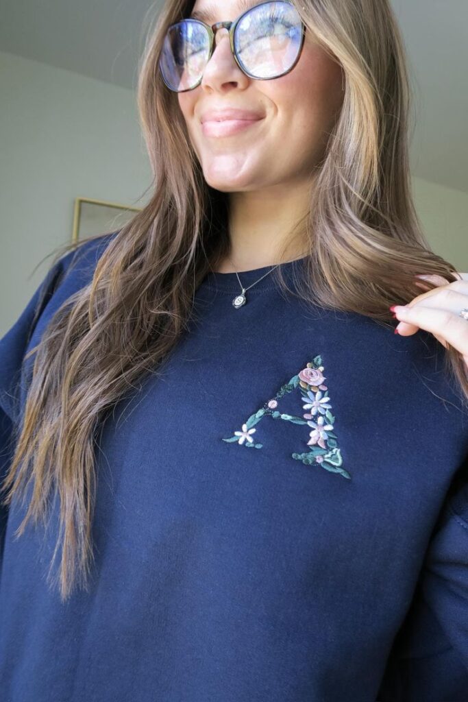 Woman in glasses wearing a navy sweater with floral embroidered A, smiling indoors.