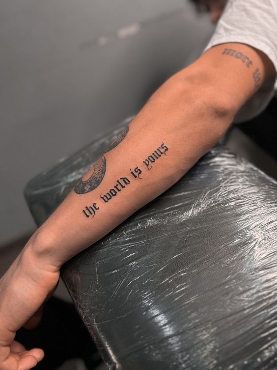Forearm tattoo with text the world is yours in black script, arm resting on covered surface.