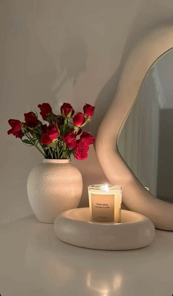 Aesthetic decor with red roses in a vase, lit candle, and modern mirror on a minimalist tabletop.