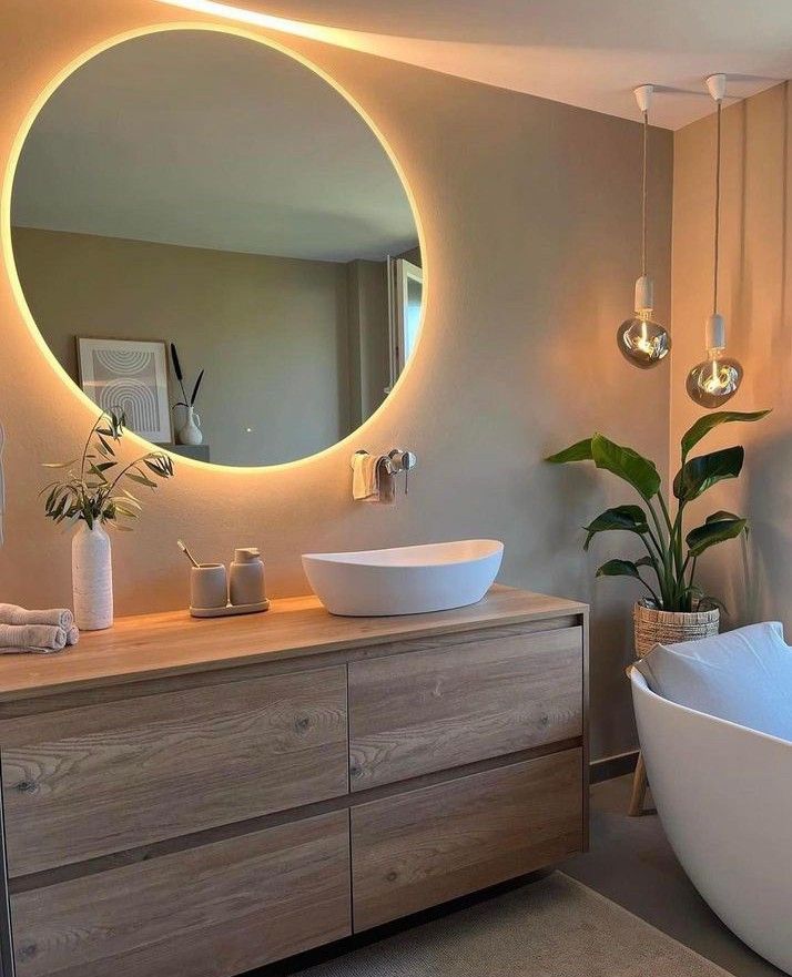 Modern bathroom with round backlit mirror, wooden vanity, vessel sink, and greenery for a serene, stylish ambiance.