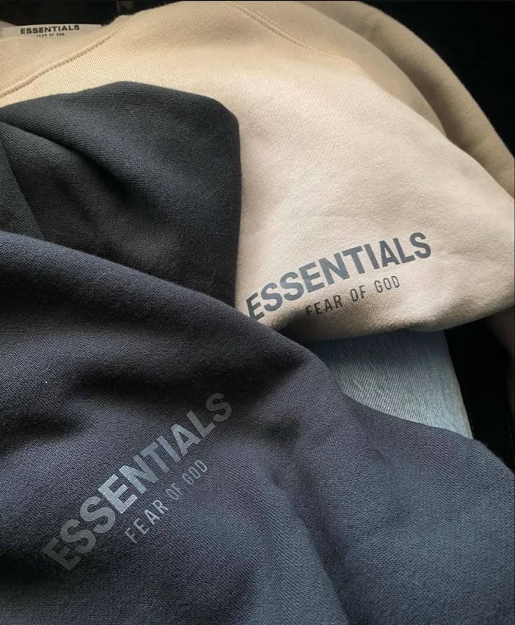 Close-up of Essentials Fear of God hoodies in beige and black, showcasing minimalist design and logo on fabric.