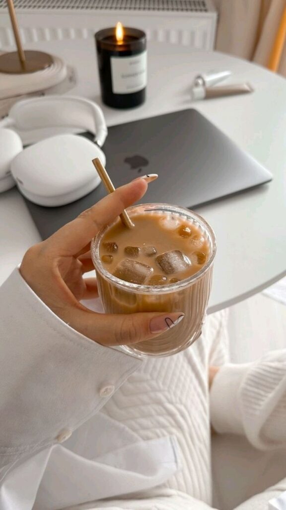 Aesthetic workspace with iced coffee, laptop, headphones, and a lit candle on a white table.