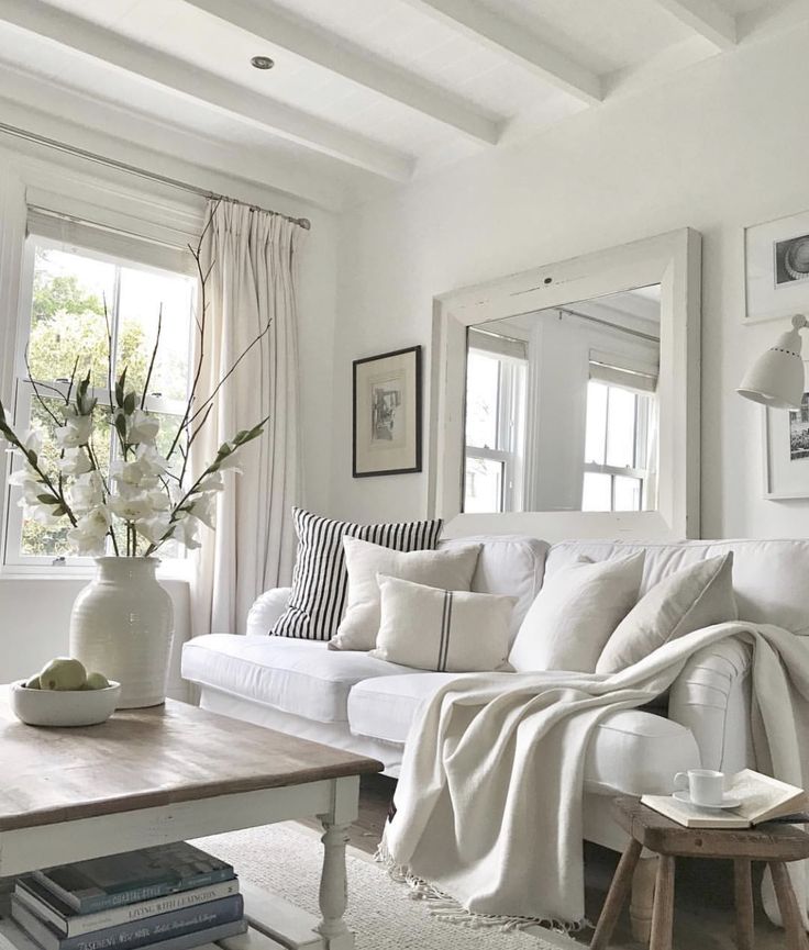 Bright, cozy living room with white sofa, decorative pillows, and large mirror. Light wood table and flowers enhance the space.