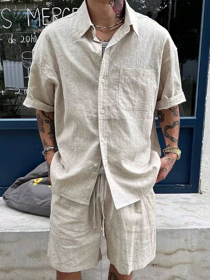 Man wearing a beige linen shirt and shorts, with tattoos and accessories, standing outdoors.