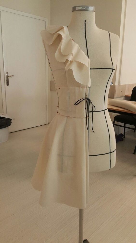 Elegant dress design on mannequin, featuring a ruffled, asymmetrical one-shoulder style in a tailoring studio.