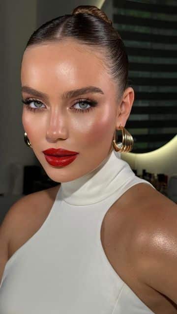 Woman with bold makeup and red lipstick wearing a white top and gold hoops, posing in a well-lit room.