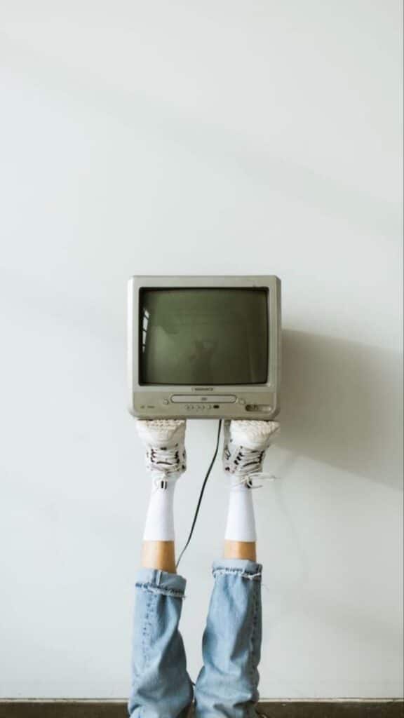 Person with TV on head and legs up, wearing jeans and sneakers against a white wall. Quirky, creative concept image.