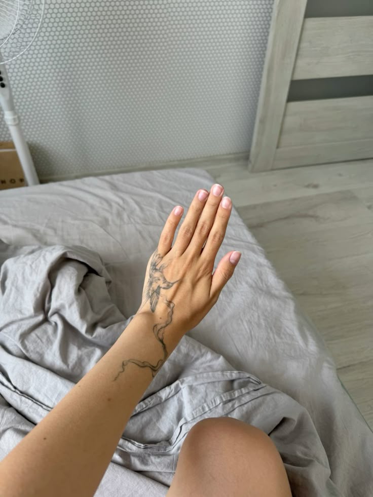 Tattooed arm with hand extended over gray bed sheets, showcasing intricate design on forearm.